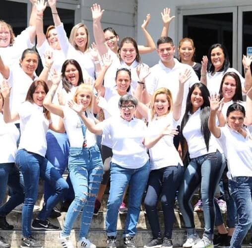 A group of 18 people standing on stairs wearing blue jeans and white shirts all with their hands up in celebration
